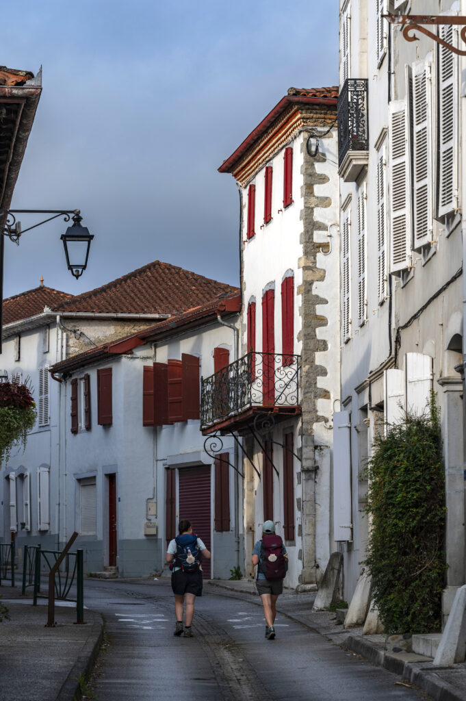 Deux pélerins avancent dans la rue du Palais de Justice à Saint-Palais. Il s'agit d'une rue du 13ème siècle, elle arbore un style d'architecture caractéristique de la Basse-Navarre. Des murs blancs et des volets rouges.